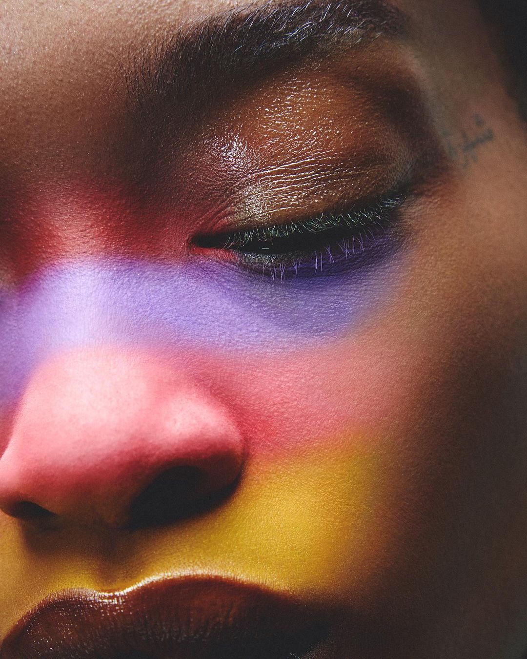Close-up studio shot of adult female face, eyes closed, with makeup in red, pink, purple, and gold tones on fair skin.