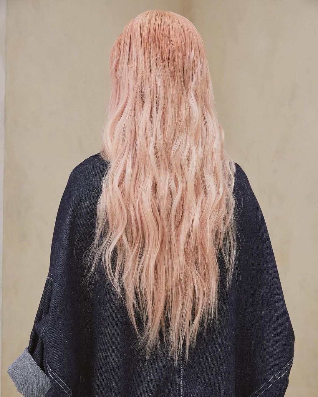 Woman with long wavy hair in shades of pink and blonde, back view, wearing a dark blue denim jacket against a tan neutral background.