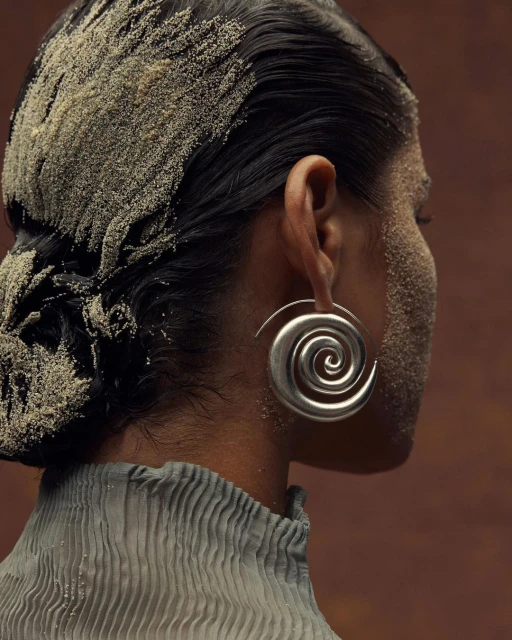 Close-up fashion shot of a woman wearing large spiral earrings, with sand scattered on her hair and face in a studio setting.