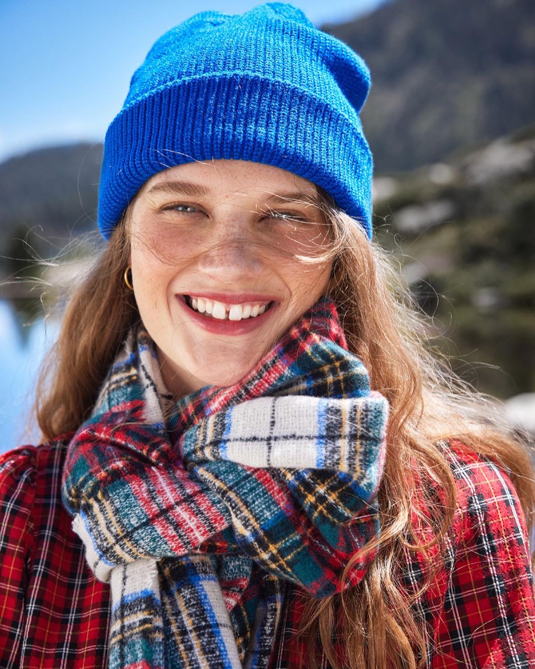 Portrait of smiling young woman wearing a blue beanie and plaid scarf, standing outside in a natural mountain environment on a sunny day.