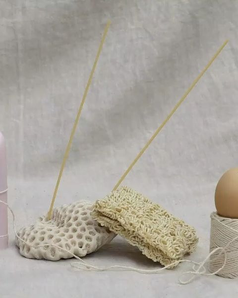 Still life of spaghetti, ramen noodles, thread spool, egg, and porous stones against a white background, showcasing textures and neutral tones.