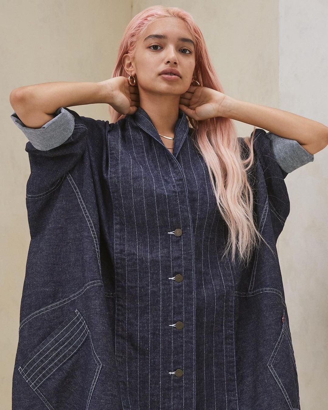 Woman with pink hair poses in a denim jacket with rolled-up sleeves and a collar, set against a neutral background in a studio setting.