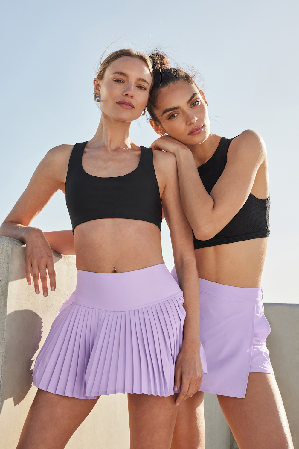 Two women in athletic wear stand against a white wall under a bright sky. One woman rests her arm on the other's shoulder.