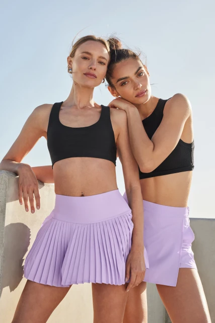 Two women in athletic wear stand against a white wall under a bright sky. One woman rests her arm on the other's shoulder.
