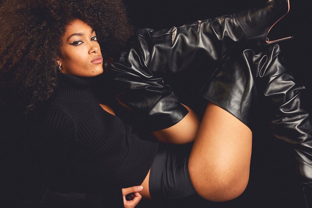 An adult female model with dark skin and large afro hairstyle poses in a black top and thigh-high black leather boots against a dark backdrop.