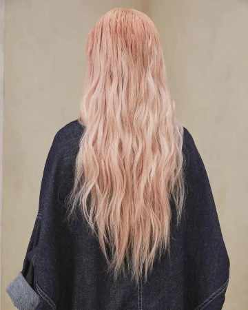 Rear view of a person with long wavy pink hair wearing a dark denim jacket against a neutral textured wall.