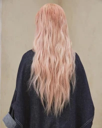 Rear view of a person with long wavy pink hair wearing a dark denim jacket against a neutral textured wall.