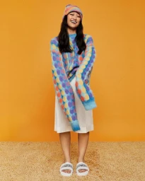 Smiling woman wearing a colourful patterned knit sweater, matching beanie, white skirt, and slides against an orange backdrop
