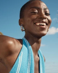 Close-up portrait of a smiling Black woman wearing a blue halter top and silver hoop earrings against a bright blue sky.