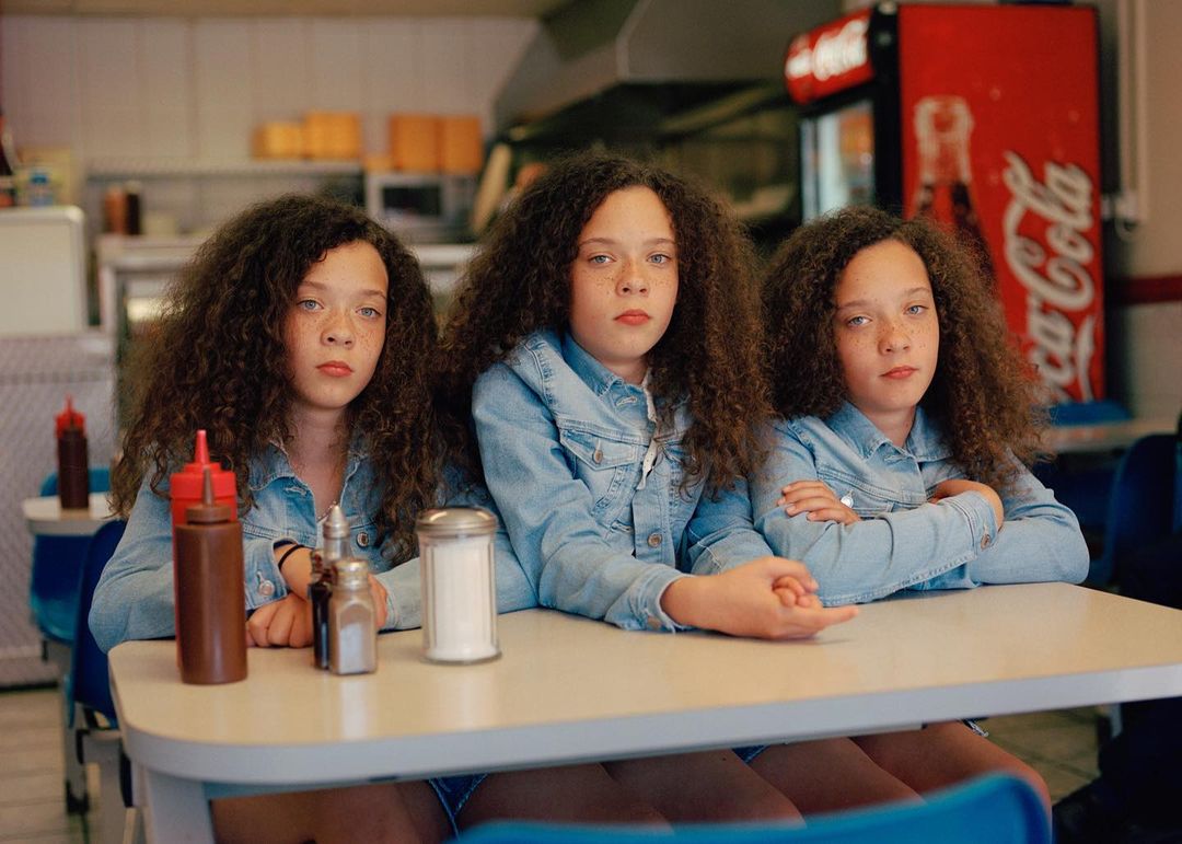 Three young women with curly hair in denim jackets sit at a table with condiments and a soda machine in the background.