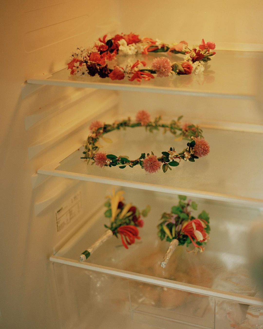 Flower crowns and floral bouquets are displayed on glass shelves inside of a white refrigerator, offering a unique, cool storage.