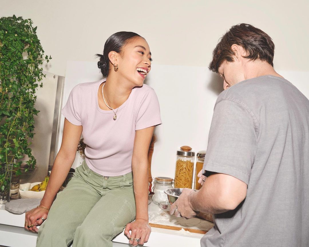 Young woman sits on counter smiling at man preparing food in kitchen. The bright and airy indoor setting includes plants.