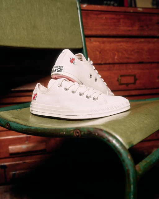 A pair of white Converse All Star sneakers with a floral design rest on a green metal chair, set against a wooden backdrop.