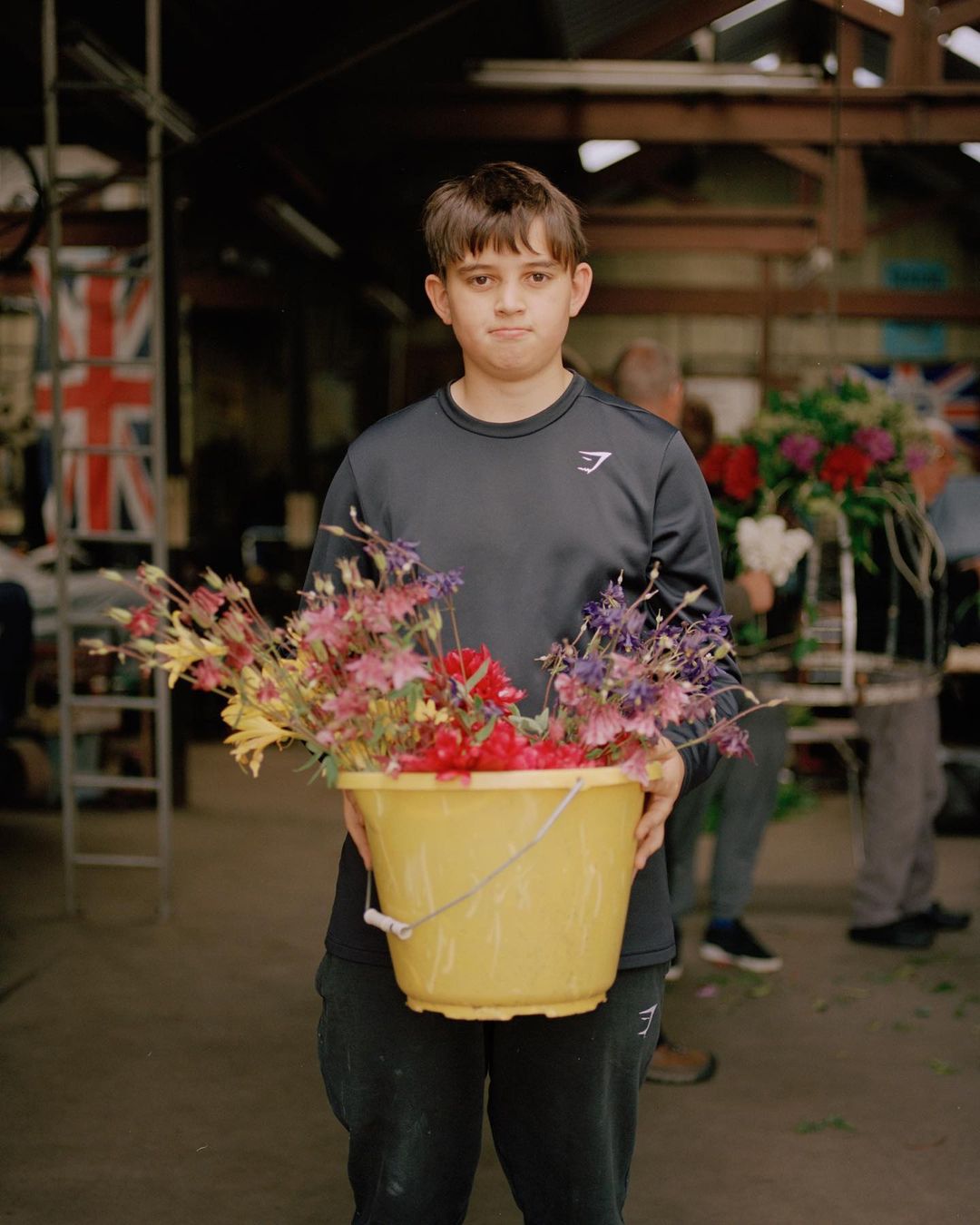 Boy in grey holds yellow bucket of cut flowers indoors with Union Jack flag and other people in background, medium shot, eye-level angle.