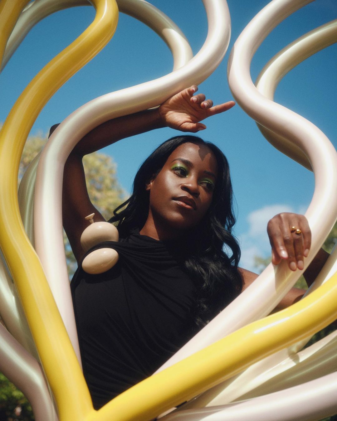 Attractive woman with bright green eyeshadow poses with art installation against a blue sky. She is wearing a black dress and gold ring.