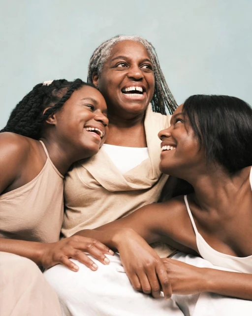 Three African American women of different generations, grandmother and two adult daughters, are laughing indoors in a natural light studio.