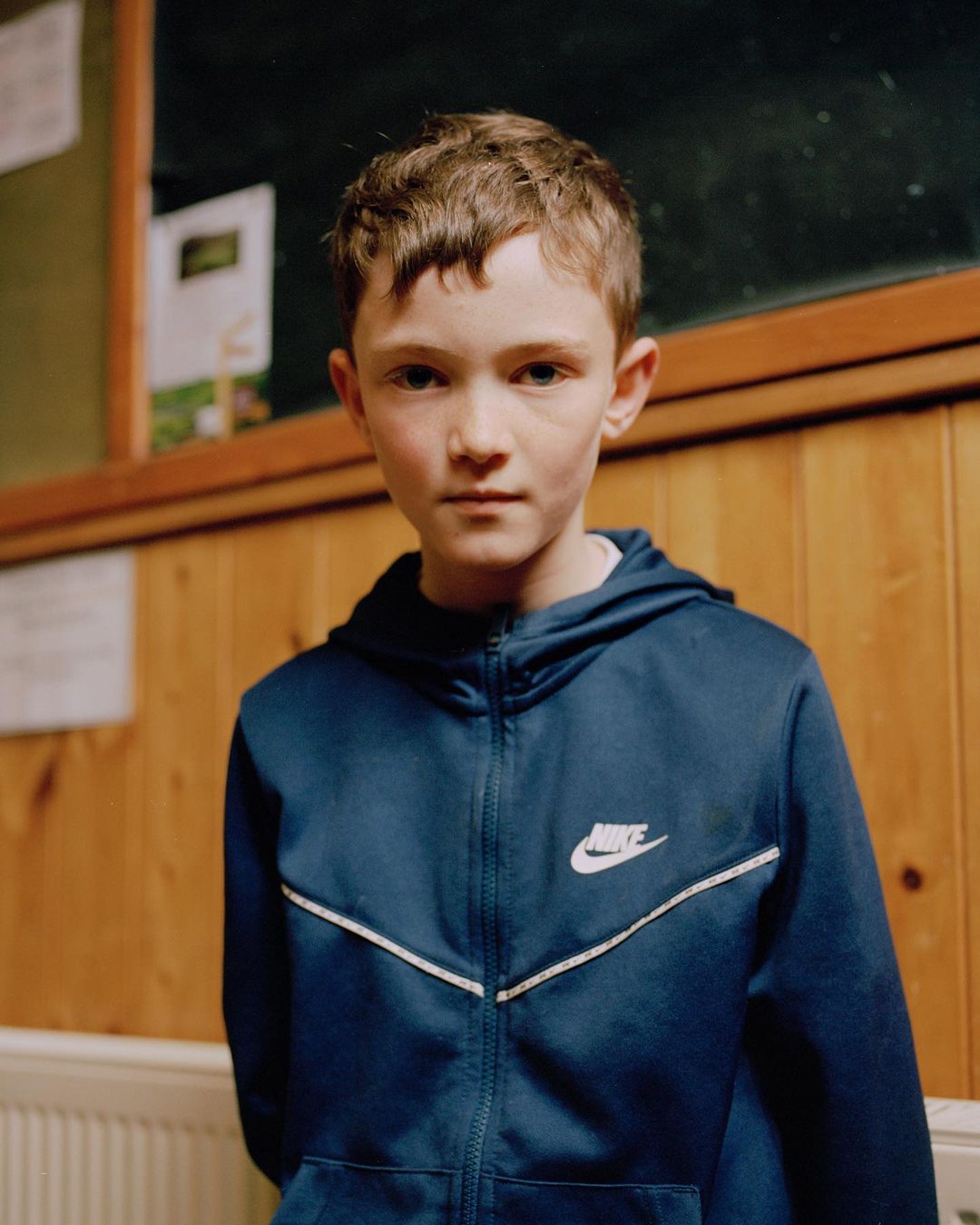 Portrait of a young boy with light brown hair wearing a blue Nike hoodie, posing indoors with wood-paneled walls and a dark green bulletin board.