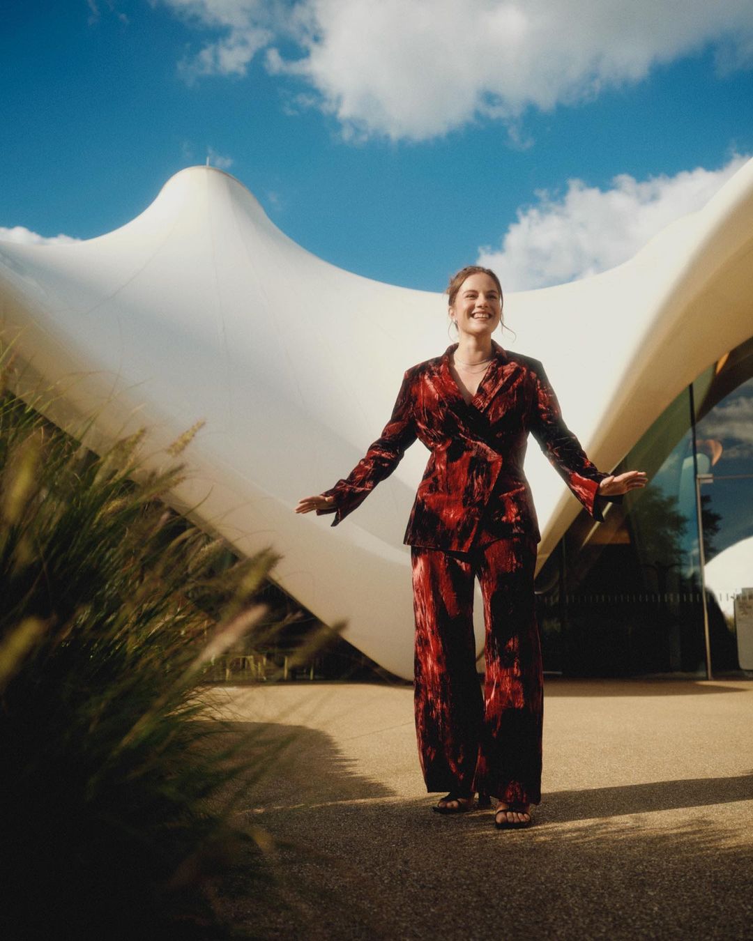 Adult woman in a patterned red suit with arms outstretched, stands on a sidewalk with blurred greenery and a white tent-like building.