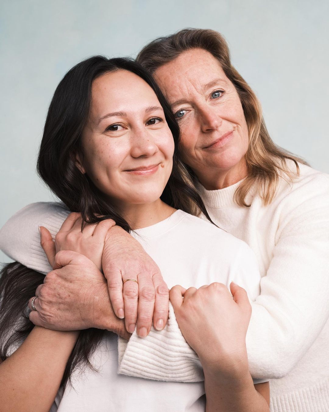 Portrait of an Asian woman embracing an older woman in a light-filled indoor photo studio, both wearing white tops, with soft, diffused lighting.
