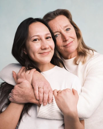 Portrait of an Asian woman embracing an older woman in a light-filled indoor photo studio, both wearing white tops, with soft, diffused lighting.