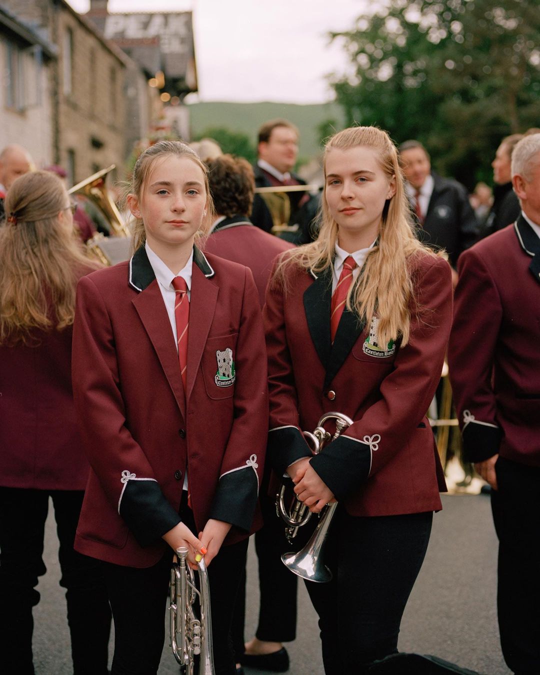 Two young female musicians in matching burgundy blazers and ties hold trumpets on a street with other musicians in the background.