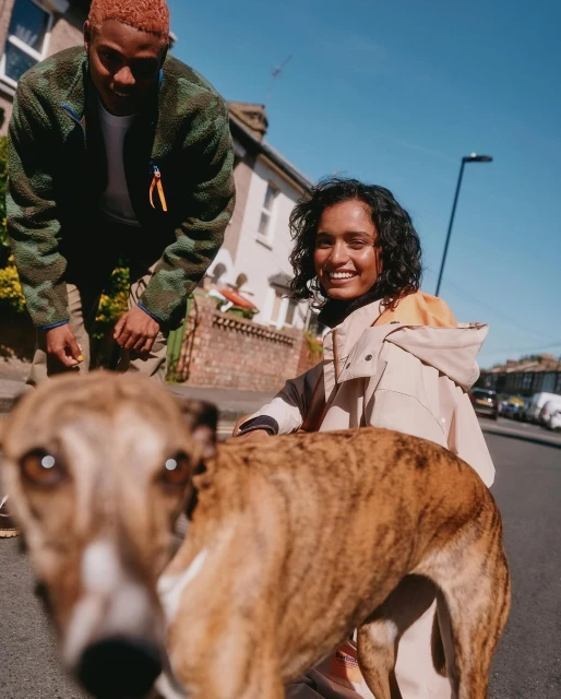 Woman smiles with her dog on a street as a man looks on in an eye-level, sunny outdoor shot in front of row houses.