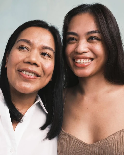 Close-up portrait of two smiling women with dark hair in a naturally lit studio. One wears a white shirt, the other a beige top.
