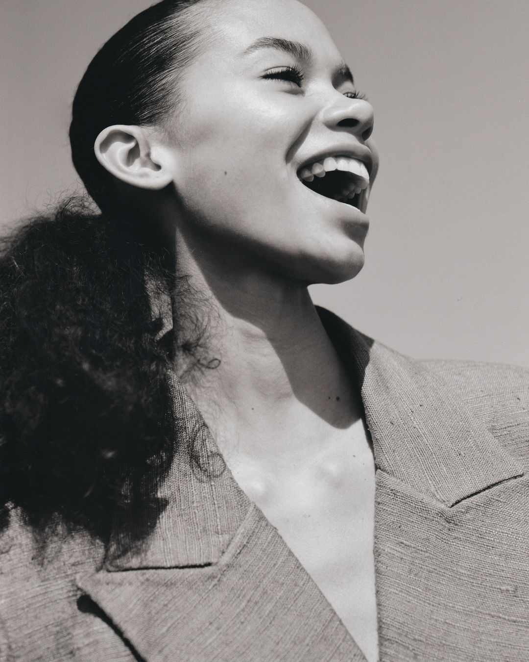 A close-up studio shot of a laughing adult woman with dark curly hair, wearing a blazer, with a clear sky background, monochrome.