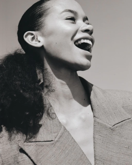 A close-up studio shot of a laughing adult woman with dark curly hair, wearing a blazer, with a clear sky background, monochrome.