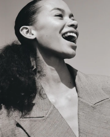 A close-up studio shot of a laughing adult woman with dark curly hair, wearing a blazer, with a clear sky background, monochrome.