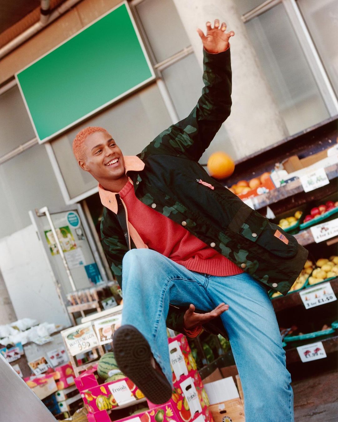 A young adult smiles and gestures while standing in front of a fruit and vegetable stand with a green sign above the store.