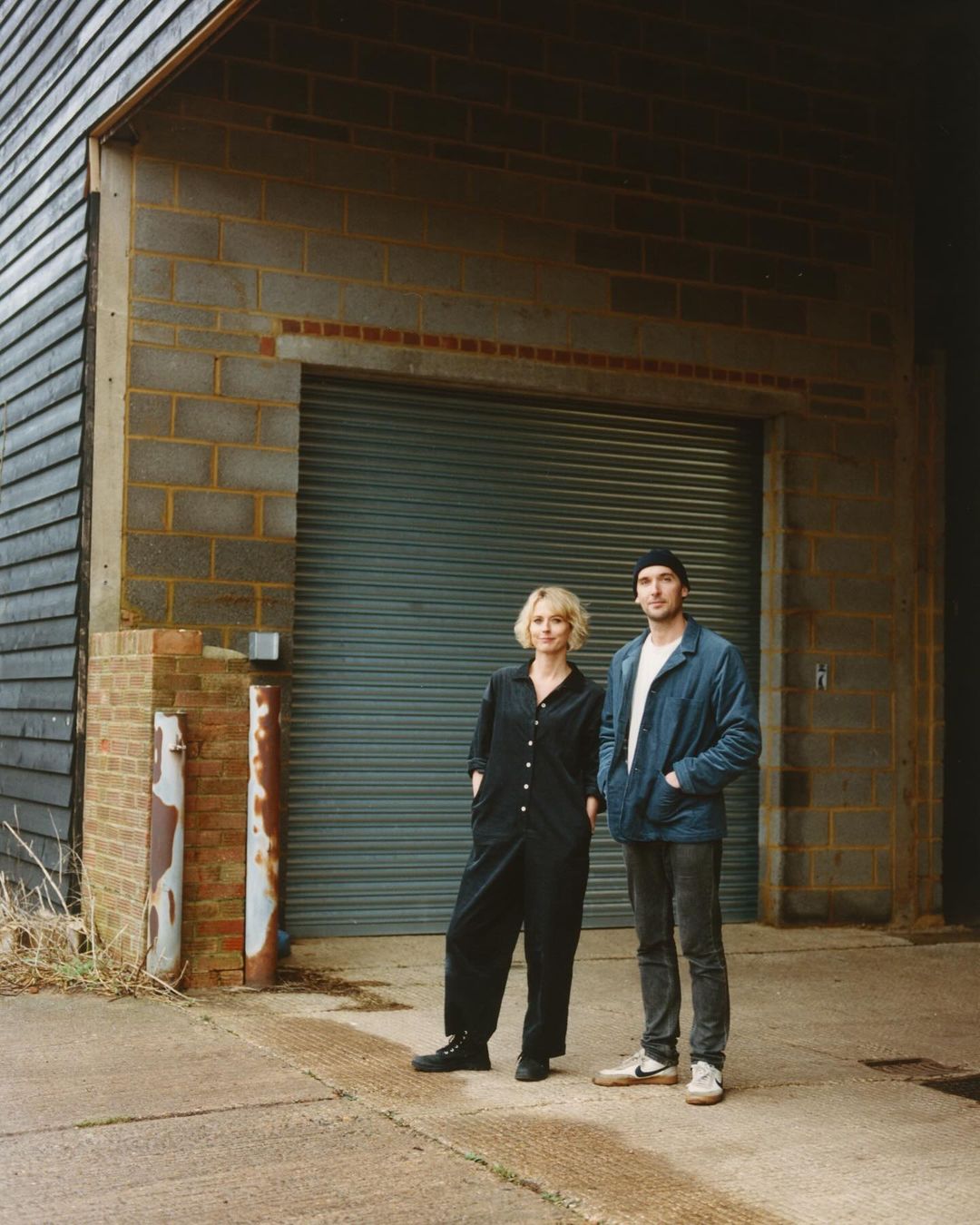 Portrait of a couple standing outdoors in front of a gray brick building with a closed roll-up door, both wearing casual clothing.