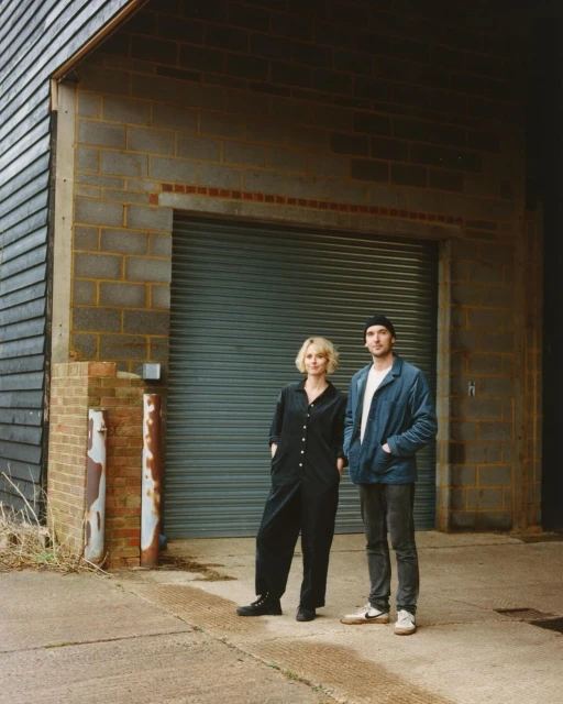 Portrait of a couple standing outdoors in front of a gray brick building with a closed roll-up door, both wearing casual clothing.