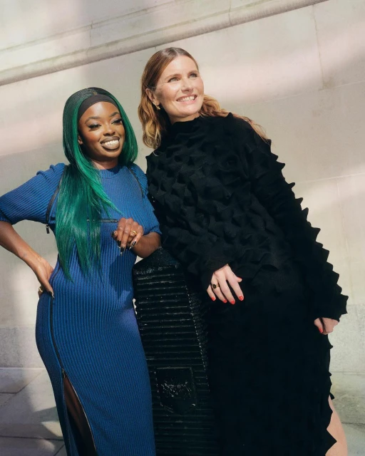 Two women pose in fashionable dresses indoors. One has teal hair and a blue dress, while the other wears a black dress.