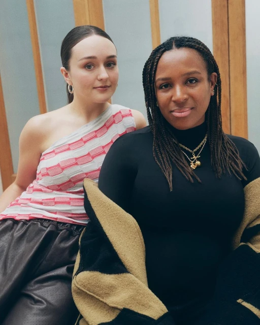 Two adult women pose indoors. One wears a pink-and-white top and dark pants, while the other wears a black turtleneck. A patterned jacket is draped over her.