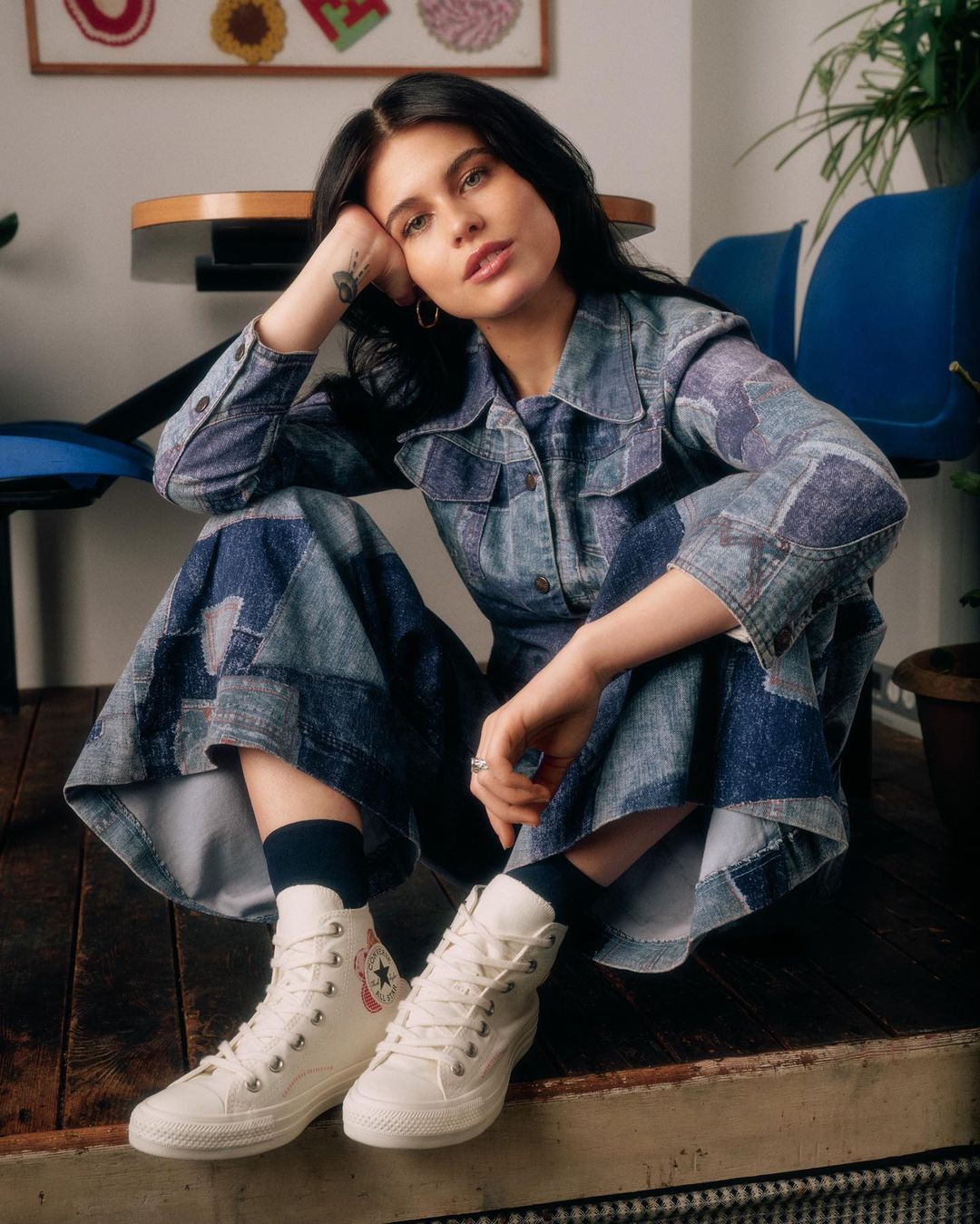 A woman with dark hair and denim dress posing indoors, seated on the floor in front of a table and blue chair wearing high-top sneakers.
