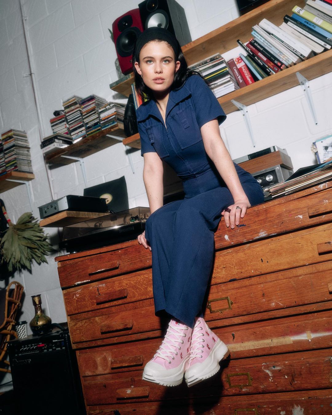 A woman in a blue jumpsuit and pink sneakers is sitting on top of a wooden dresser, inside of a room with shelves and speakers.