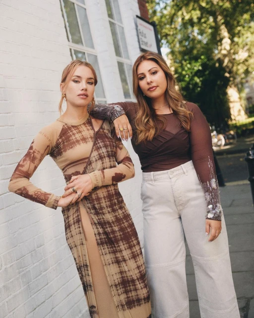 Two women pose outdoors. One in a brown pattern dress, the other in a brown top and white pants, standing by a white brick building.