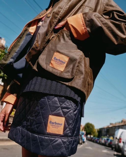 Close-up of a person outdoors wearing a Barbour jacket and quilted skirt with street and parked cars in the background.