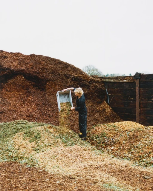 Woman unloads container of shredded material near a large mound of brown and green wood chips under a cloudy sky outdoors.