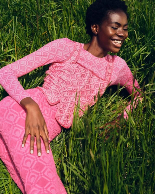 An African-American woman smiles lying in the grass wearing a pink patterned long-sleeve shirt and pants, outdoor fashion photography.