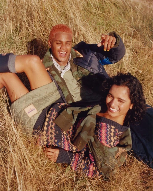 Two smiling people, an adult male and adult female, lying in golden tall grass. The couple wears quilted jackets and sweaters in an outdoor field setting.