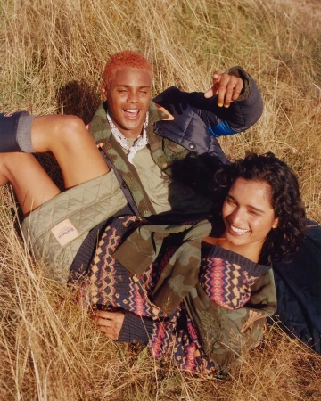 Two smiling people, an adult male and adult female, lying in golden tall grass. The couple wears quilted jackets and sweaters in an outdoor field setting.