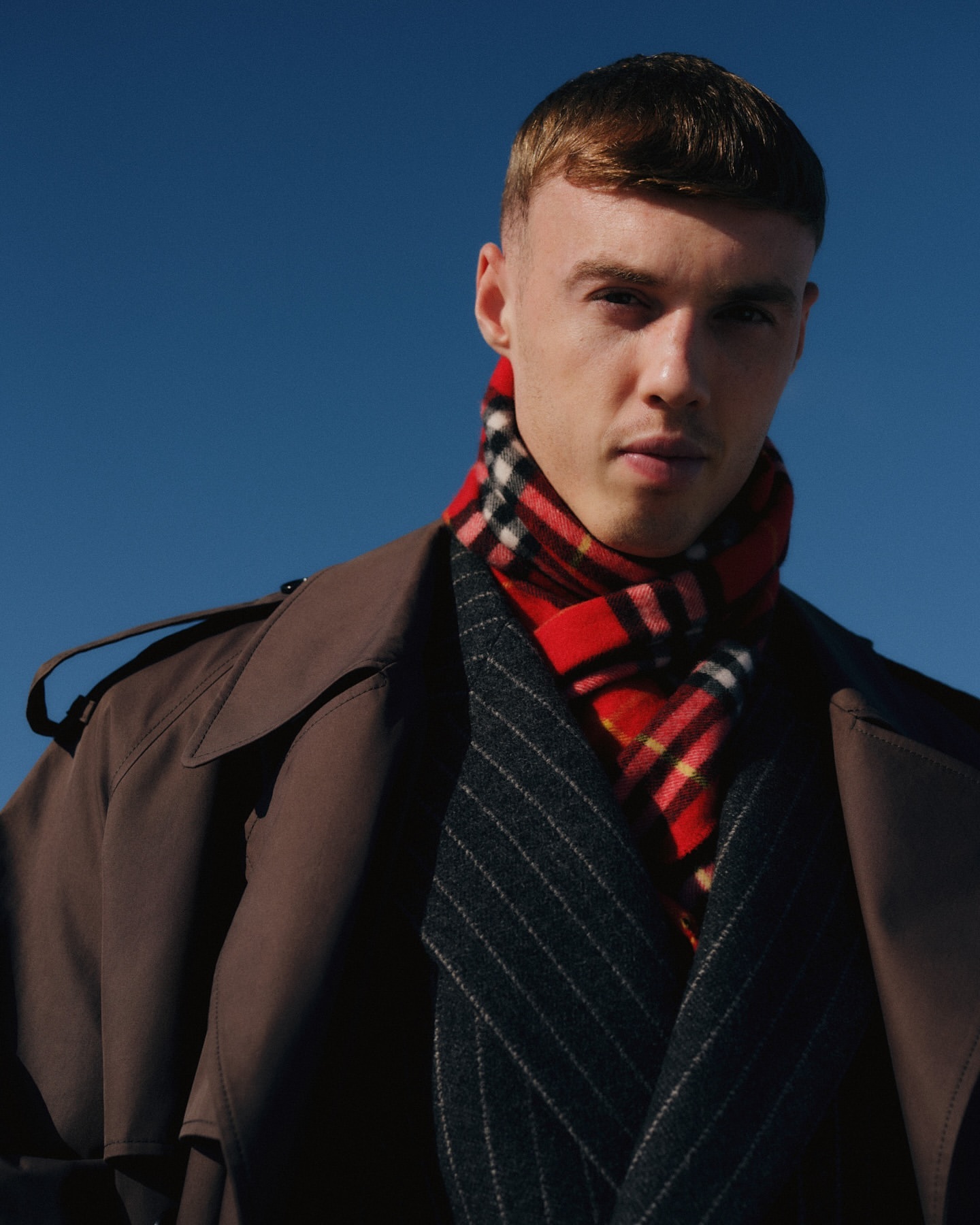 Man in a brown trench coat and a Burberry scarf against a blue sky, looking at the camera in an outdoor fashion shot.