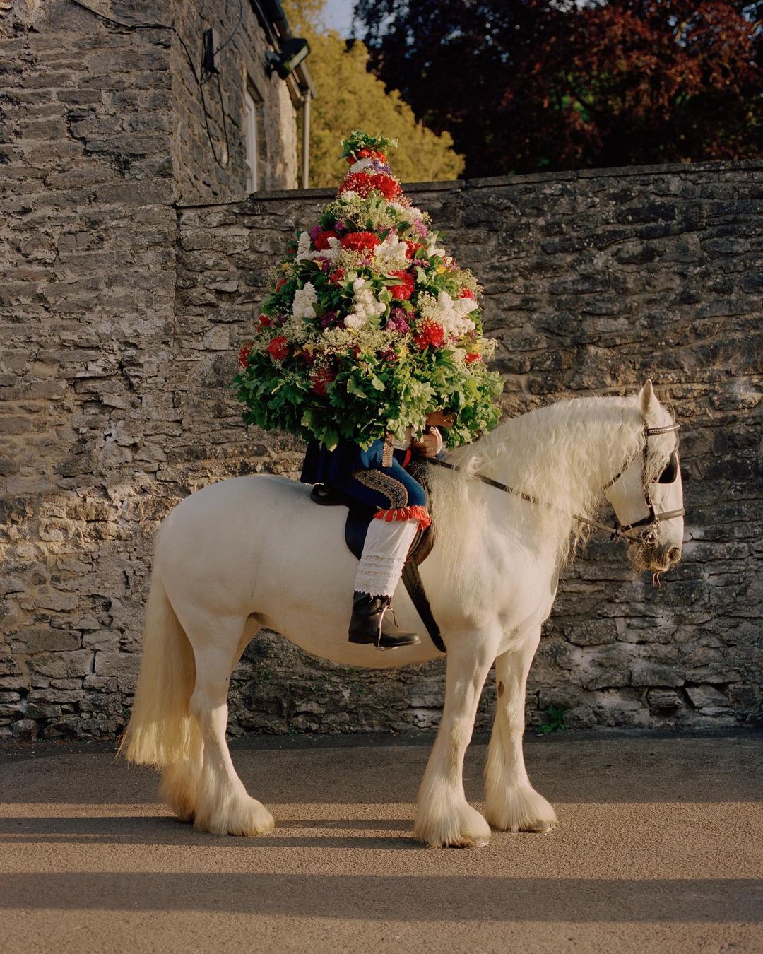 White horse adorned with floral arrangement on its back, ridden by a figure in traditional clothing, set against a stone building backdrop.