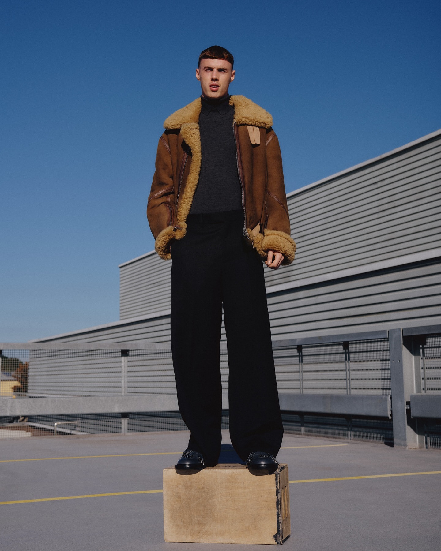 Full-length shot of a young adult man wearing a brown shearling jacket, black pants, and black shoes, standing on a concrete surface.