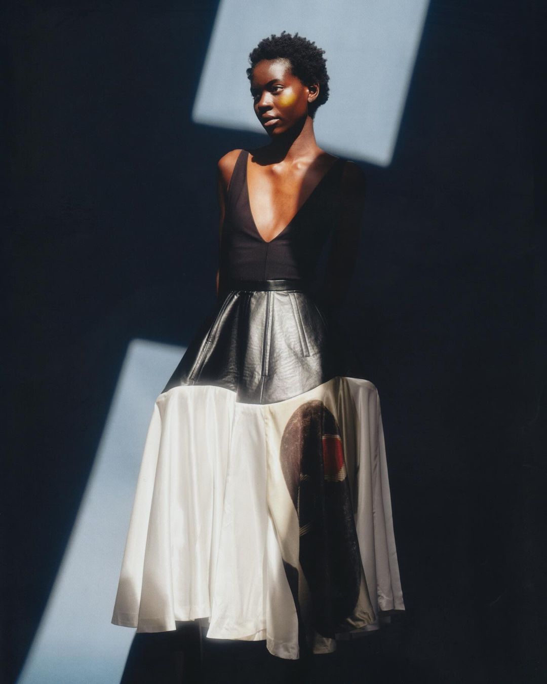 Fashion editorial shot of a black woman in a black top and graphic skirt, posing indoors under artificial light.