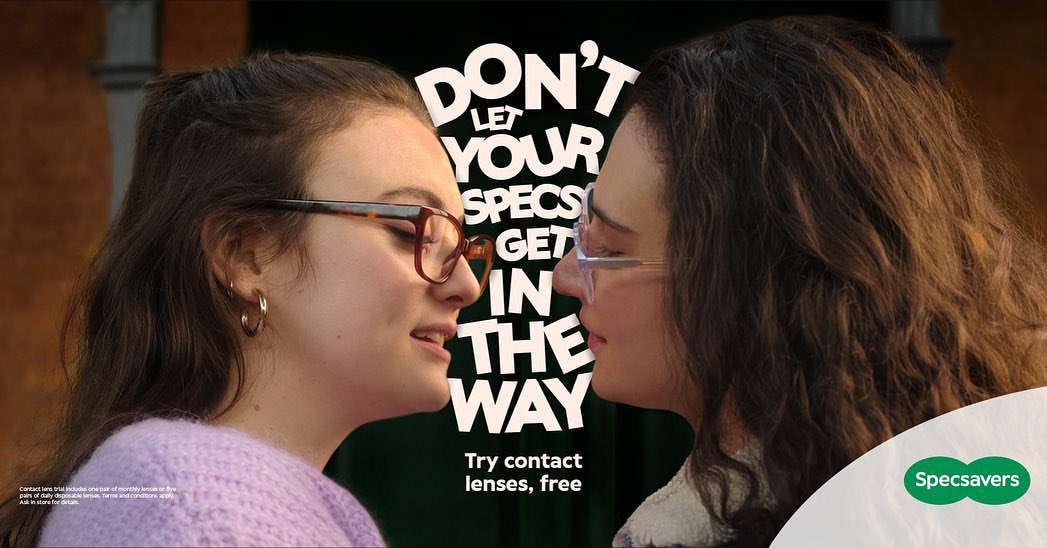 Close-up studio shot of two young adult women, faces near each other, wearing glasses, with a Specsavers ad and dark backdrop.