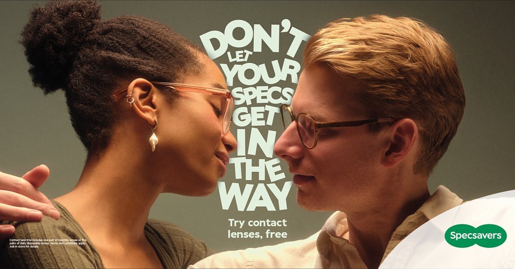 An eye-level, close-up shot features two people wearing glasses, positioned closely together, against a grey backdrop with text and a Specsavers logo.