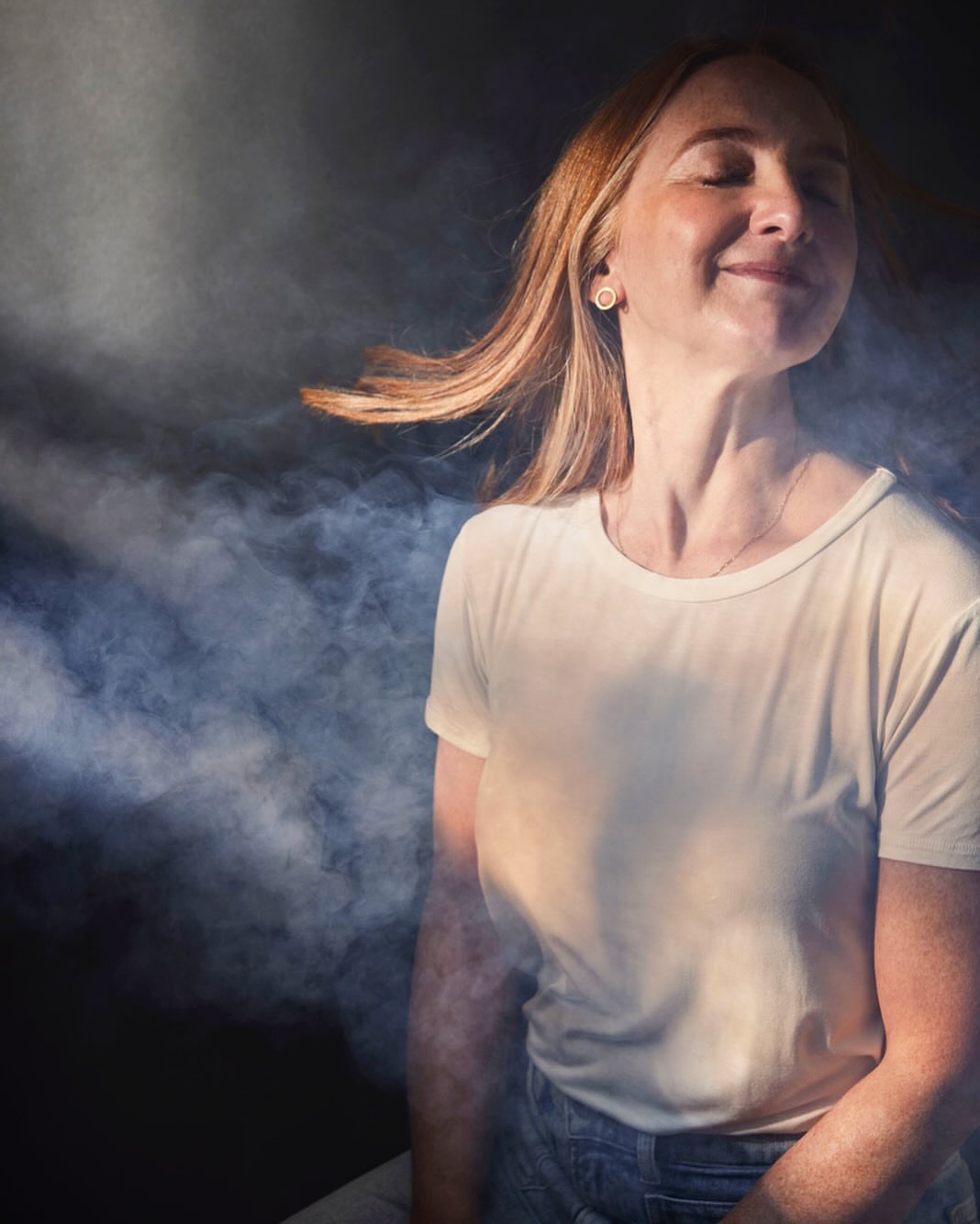 Portrait of a smiling adult woman with auburn hair and a white t-shirt posing in front of a smokey dark background with sunlight.